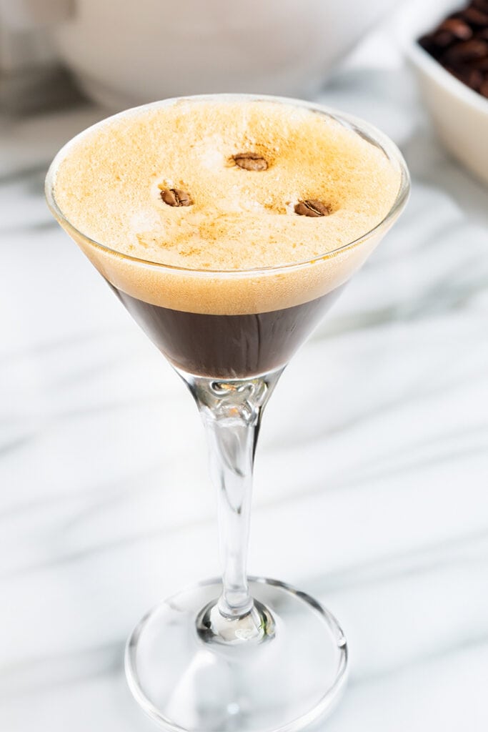 A close up image of a Espresso Martini topped with a layer of foam and coffee beans in a clear martini glass.