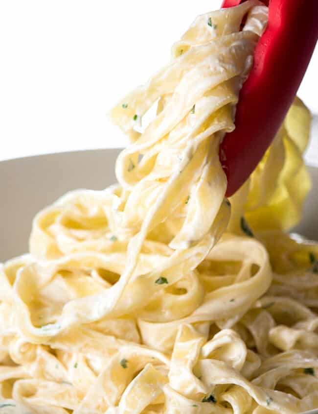 A close up image of pair of cooking tongs serving a portion of Fettuccine Alfredo into a bowl.