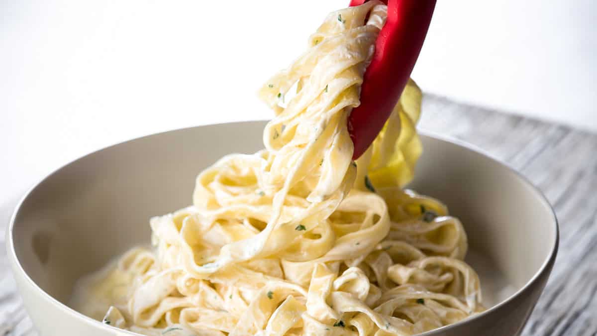 A close up image of pair of cooking tongs serving a portion of Fettuccine Alfredo into a bowl.