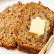 two slices of zucchini bread with a pat of melting butter on top, served on a white plate with a gold fork to the side.