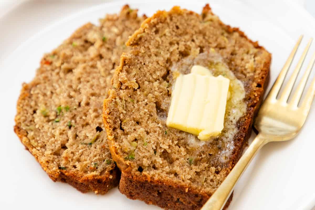 two slices of zucchini bread with a pat of melting butter on top, served on a white plate with a gold fork to the side.