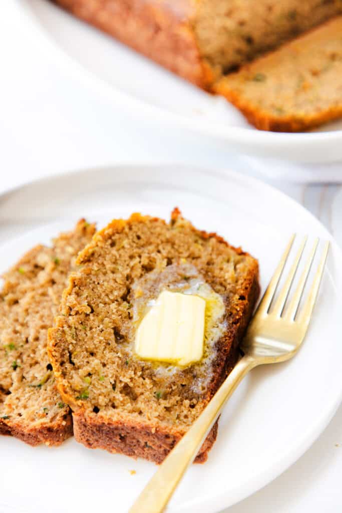 A slice of zucchini bread with a pat of melting butter on top, served on a white plate with a gold fork to the side.