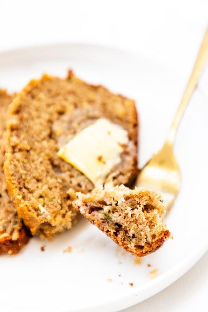 A slice of homemade zucchini bread with a bite taken out, topped with a pat of butter on a white plate with a gold fork in the background.