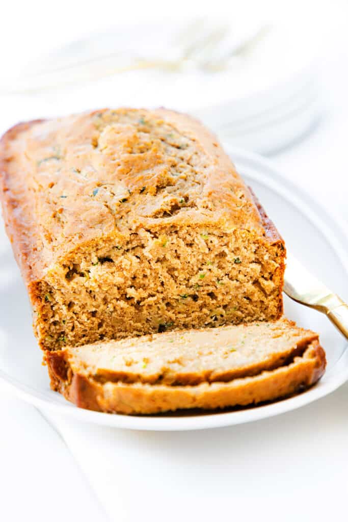 A loaf of zucchini bread with a golden-brown crust, resting on a white plate with a gold fork on the side, and slices revealing a moist, dense interior with visible bits of grated zucchini.