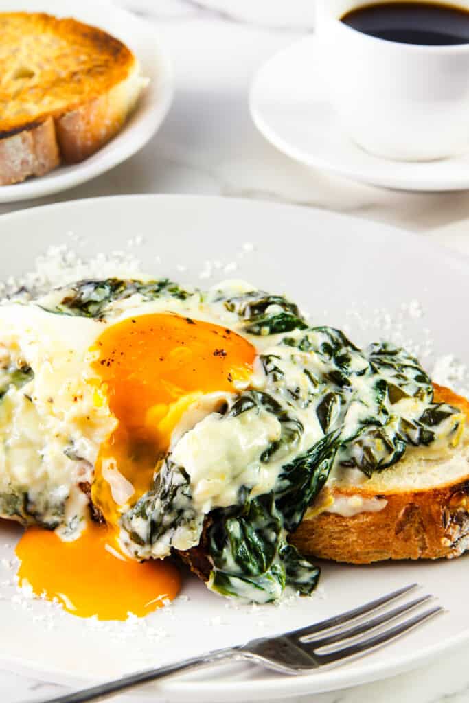A close-up of a plated serving of Baked Eggs Florentine. It's beautifully presented with a runny egg yolk spilling over creamy spinach and melted cheese, all sitting on a slice of toasted bread.