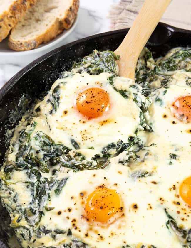 A skillet with Baked Eggs Florentine. This dish features eggs with runny yolks atop a creamy bed of spinach and melted cheese, lightly seasoned with black pepper. Beside the skillet, there’s a piece of toasted bread. The preparation appears to be fresh from the oven, ready to be served.