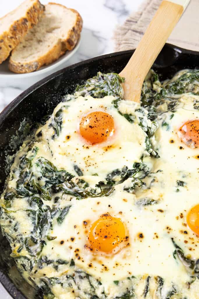 A skillet with Baked Eggs Florentine. This dish features eggs with runny yolks atop a creamy bed of spinach and melted cheese, lightly seasoned with black pepper. Beside the skillet, there’s a piece of toasted bread. The preparation appears to be fresh from the oven, ready to be served.