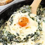 The photo displays Baked Eggs Florentine up close. It showcases eggs baked in a skillet, with one egg's yolk front and center, bright and slightly runny. The eggs are embedded in a rich mix of melted cheese and creamy spinach.