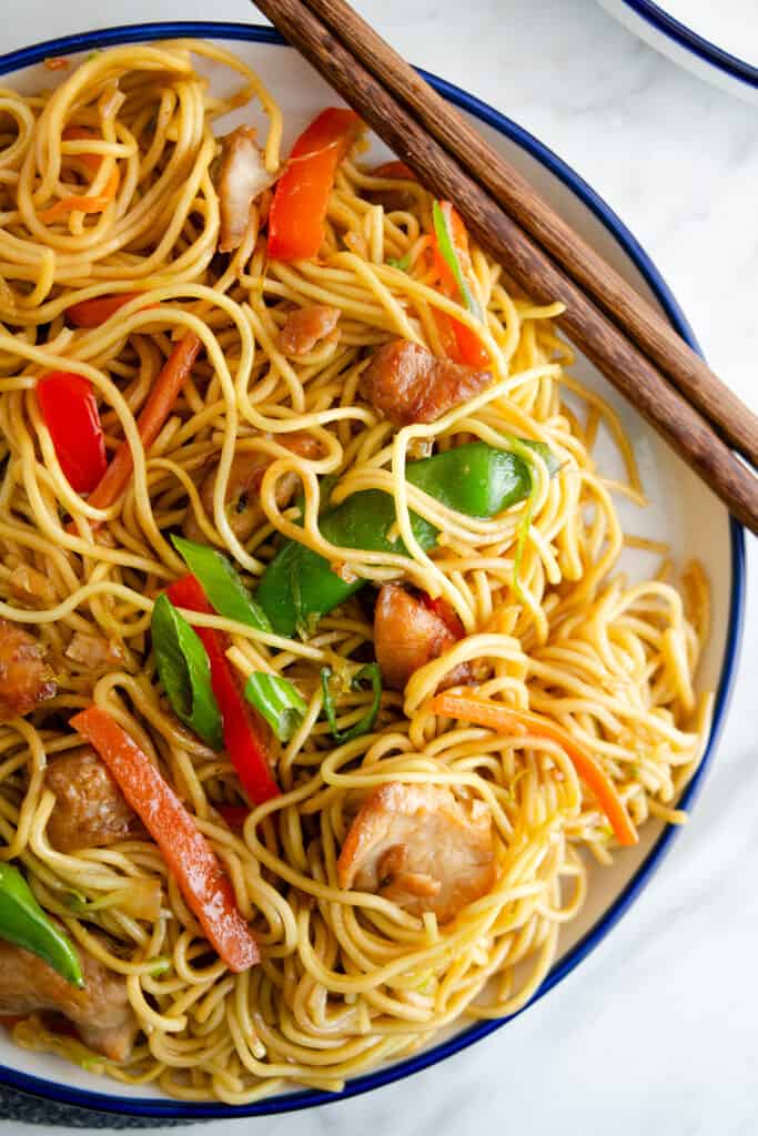 A bowl of chicken lo mein with stir-fried noodles, sliced chicken, bell peppers, carrots, and green onions, served with chopsticks on the side.