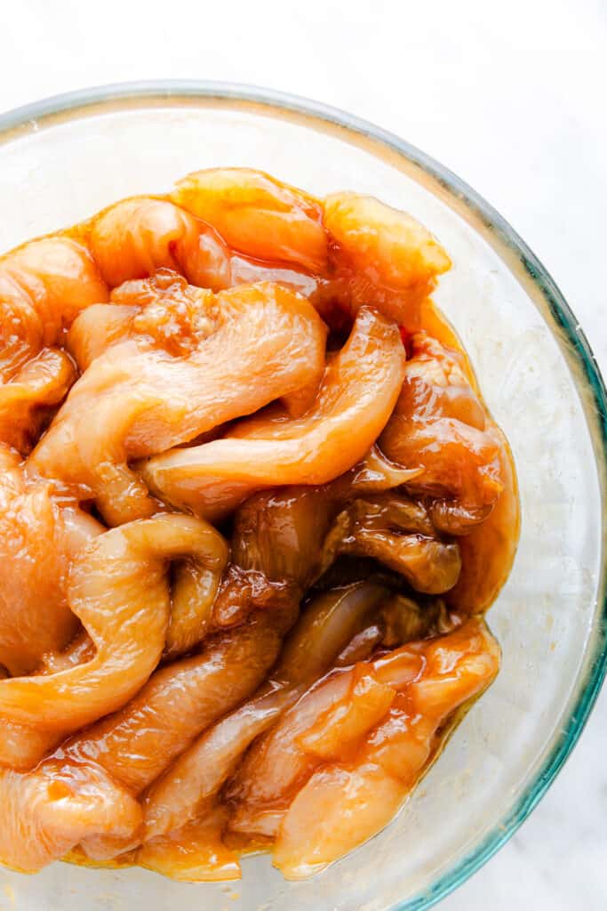 A glass bowl containing raw chicken strips marinated in a glossy amber sauce.