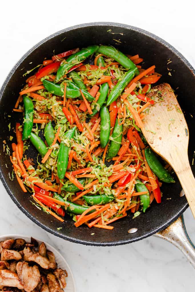 Stir-fried vegetables in a skillet with a wooden spoon, next to a plate of cooked chicken, on a marble countertop.