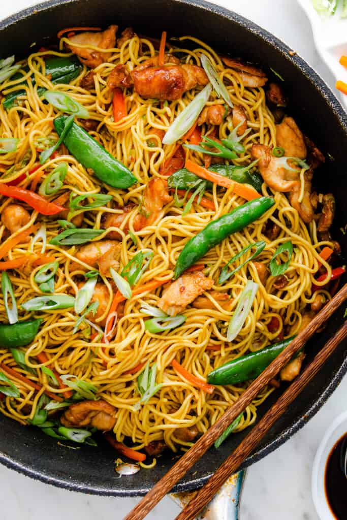 Chicken lo mein in a skillet with noodles, vegetables, and snap peas, garnished with green onions, on a marble surface.