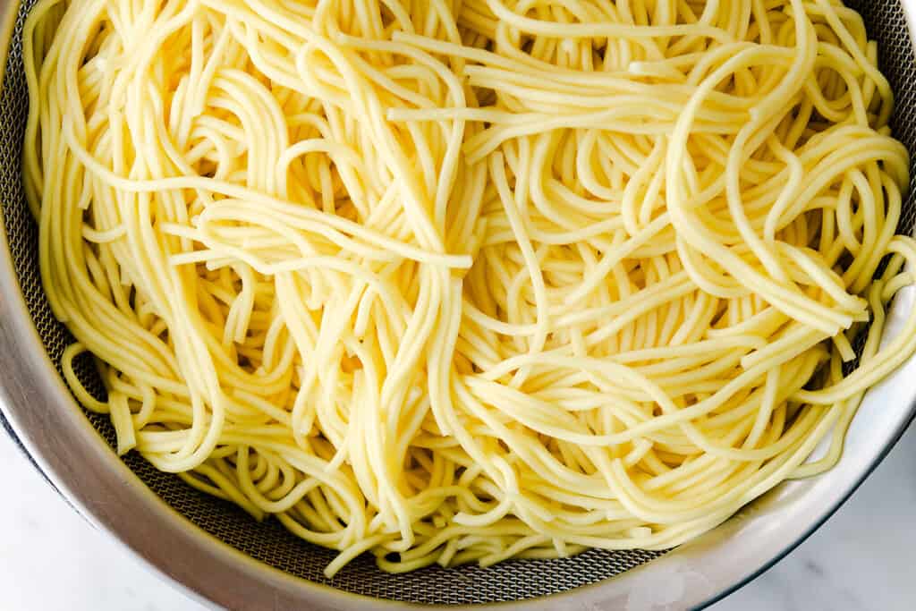 A strainer full of cooked lo mein noodles on a marble surface.