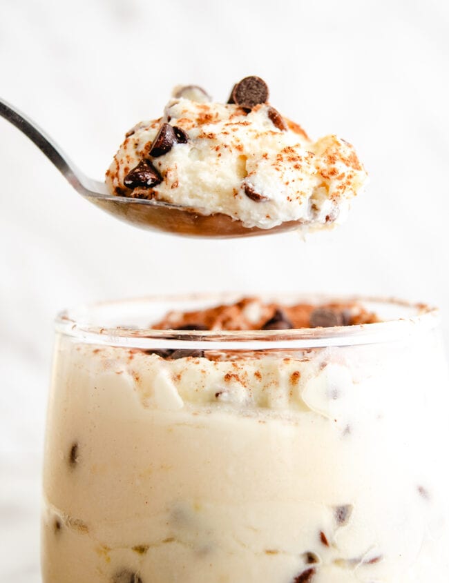 A spoon lifting a portion of creamy ricotta dessert sprinkled with chocolate chips and a dash of cinnamon, with more of the dessert visible in the glass below