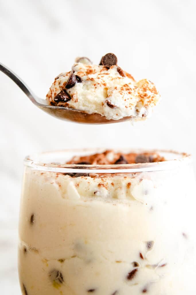 A spoon lifting a portion of creamy ricotta dessert sprinkled with chocolate chips and a dash of cinnamon, with more of the dessert visible in the glass below