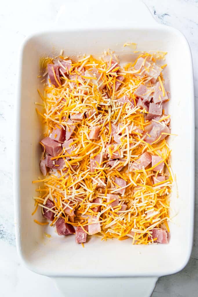 Diced ham and shredded cheddar cheese in a white baking dish, prepared for making a casserole.