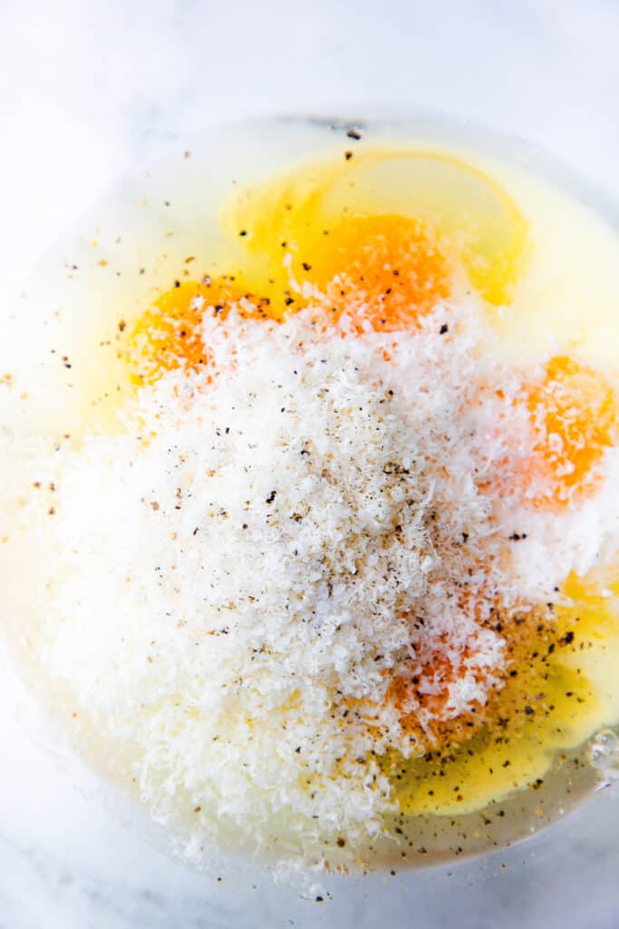 A close-up of raw eggs in a bowl with grated cheese and seasoning on top, ready for mixing.