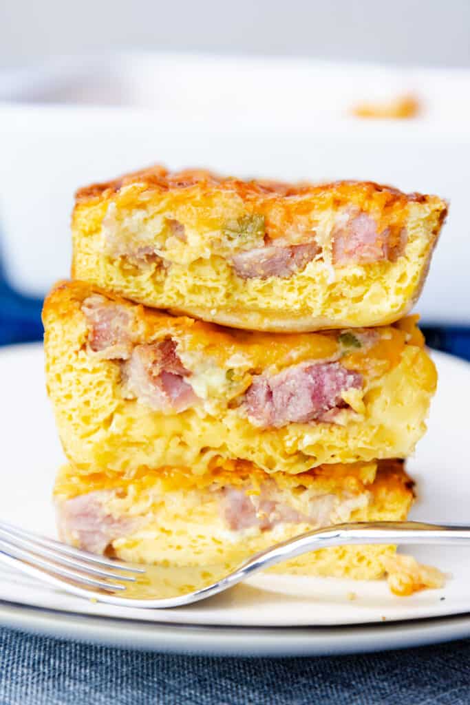 Three stacked slices of ham and egg casserole with melted cheese on a white plate, with a fork to the side.