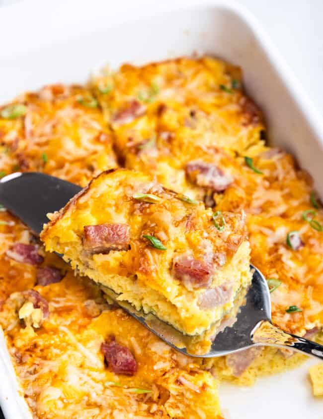 A slice of ham and cheese casserole being lifted with a spatula, showing the layered interior, garnished with herbs.