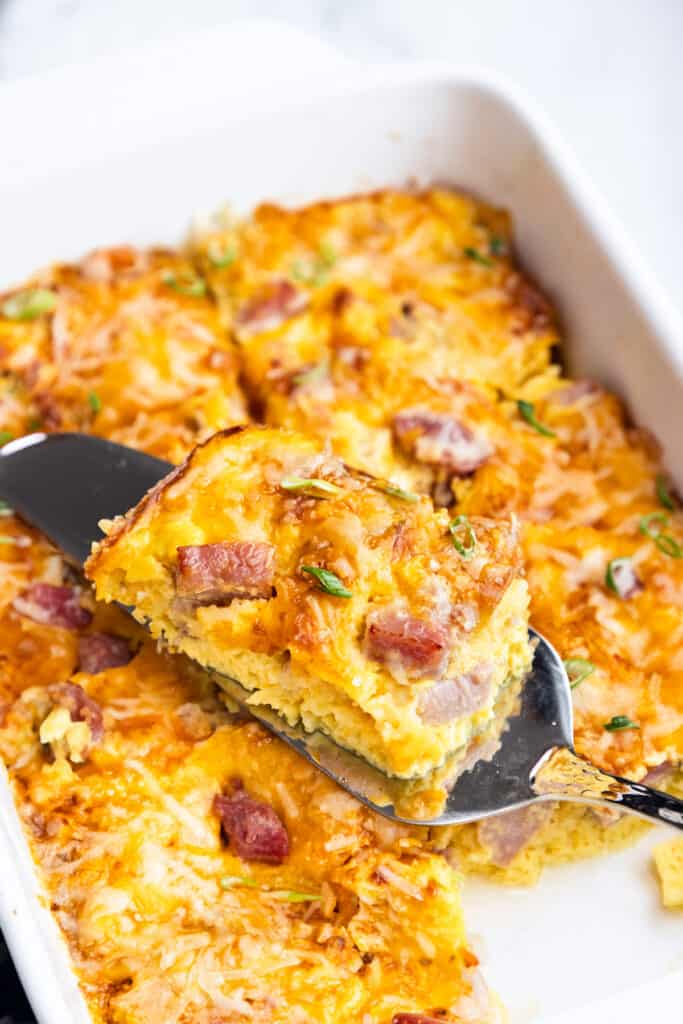 A slice of ham and cheese casserole being lifted with a spatula, showing the layered interior, garnished with herbs.