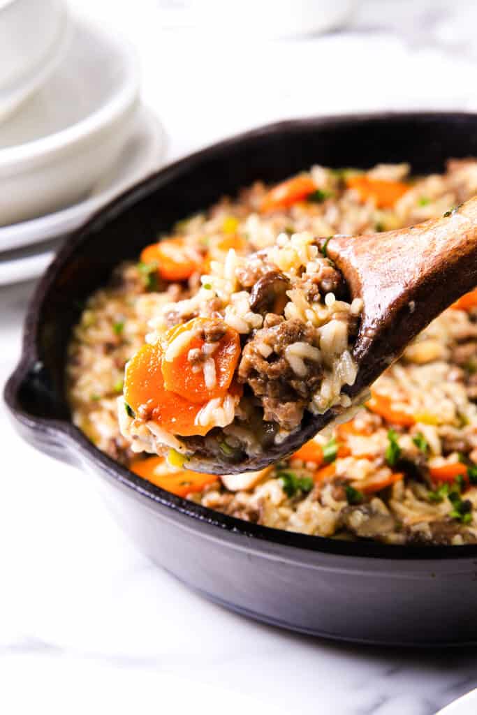 A wooden spoon scooping a portion of hamburger and rice with visible carrots and ground beef from a cast-iron pan.