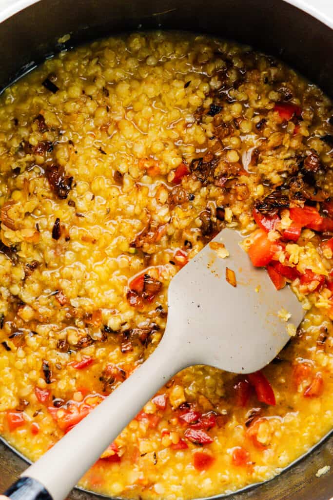 A pan of simmering red lentil dal with tomatoes and onions, stirred by a white spatula.