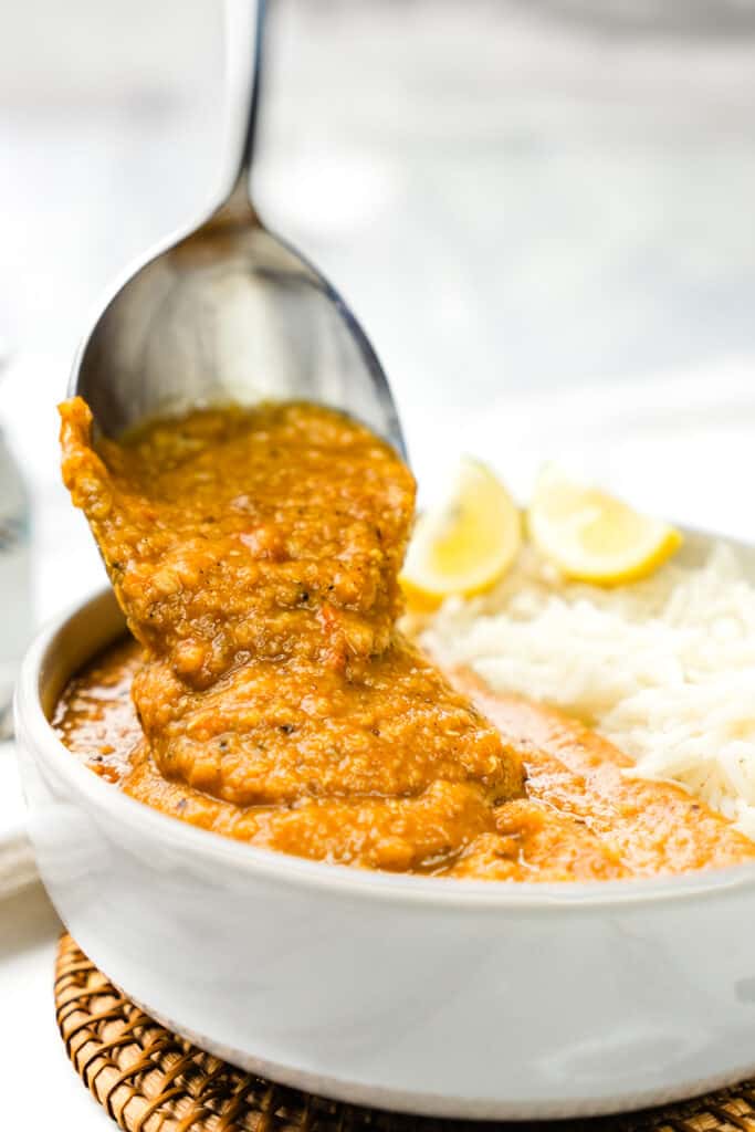 A spoon scooping up vibrant Red Lentil Dahl from a bowl, with fluffy white rice and lemon wedges in the background, all resting on a woven mat.