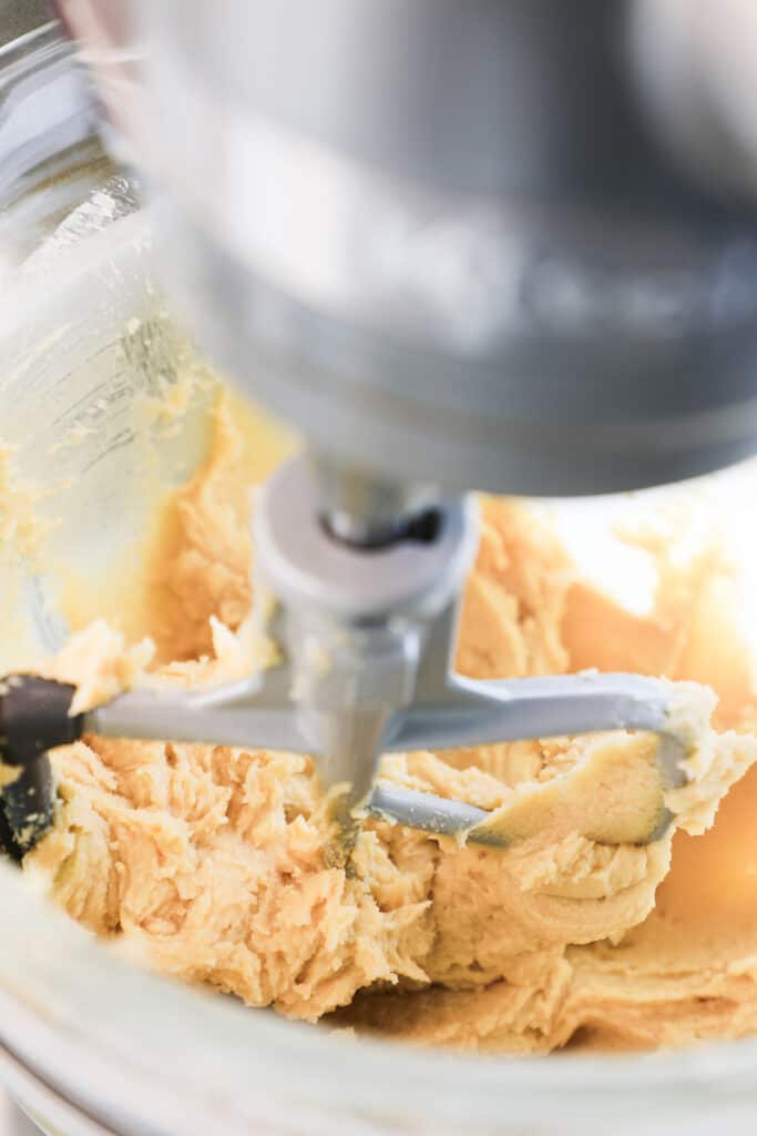 A close-up of cookie dough being mixed in a stand mixer with a paddle attachment, showing the creamy texture of the batter in a clear glass bowl.