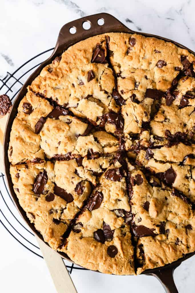 A freshly baked skillet cookie with gooey chocolate chunks, presented in a cast-iron pan on a wire cooling rack, with a wooden spatula.