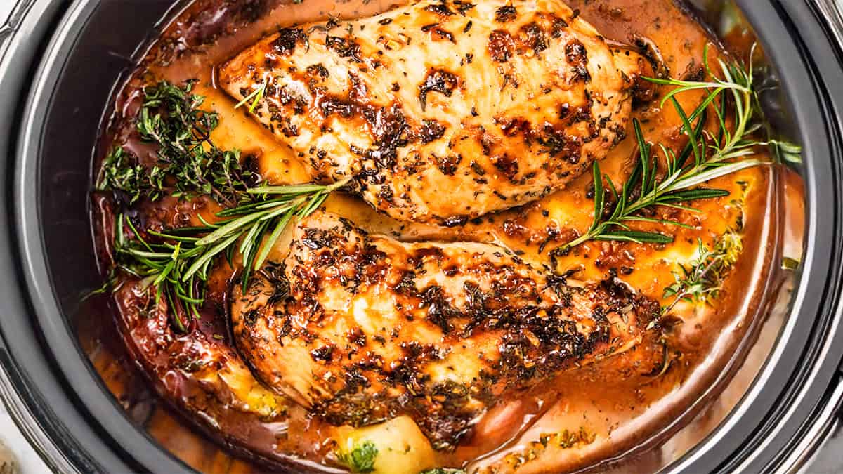 Close-up of turkey tenderloin and seasoned chicken breasts cooked in a slow cooker with herbs, rosemary, and vegetables in a savory sauce.