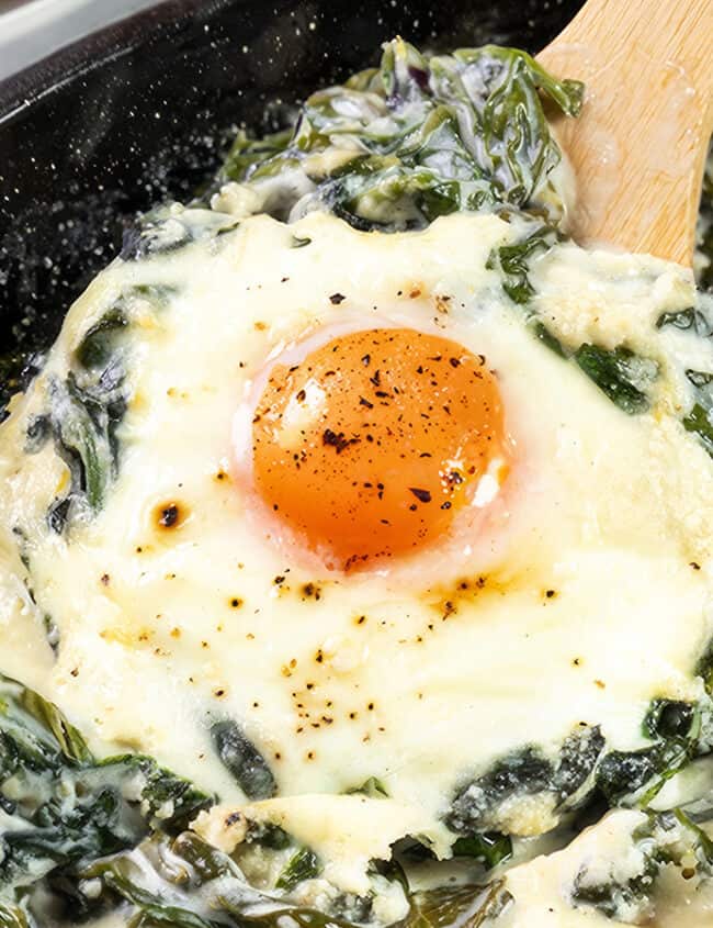 The photo displays Baked Eggs Florentine up close. It showcases eggs baked in a skillet, with one egg's yolk front and center, bright and slightly runny. The eggs are embedded in a rich mix of melted cheese and creamy spinach.