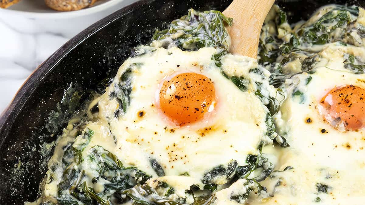 The photo displays Baked Eggs Florentine up close. It showcases eggs baked in a skillet, with one egg's yolk front and center, bright and slightly runny. The eggs are embedded in a rich mix of melted cheese and creamy spinach.