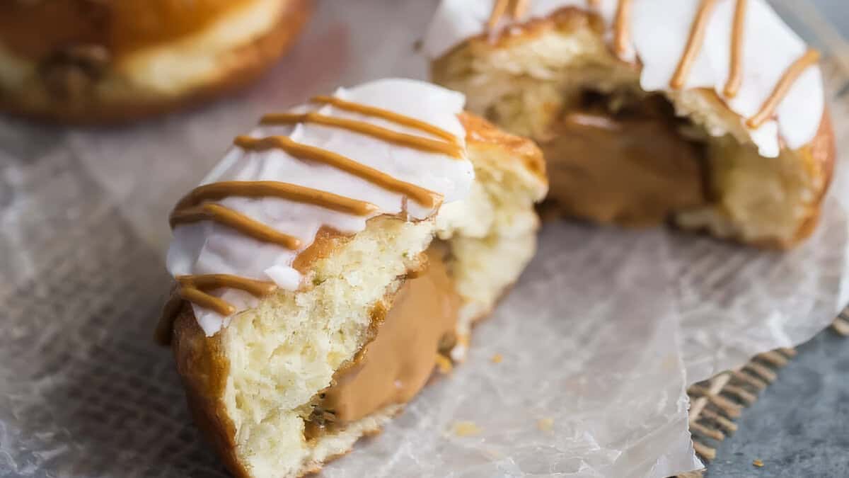 A close-up photo of a glazed biscoff donut with a peanut butter filling cut in half on a white plate. There are other whole biscoff donuts in the background.