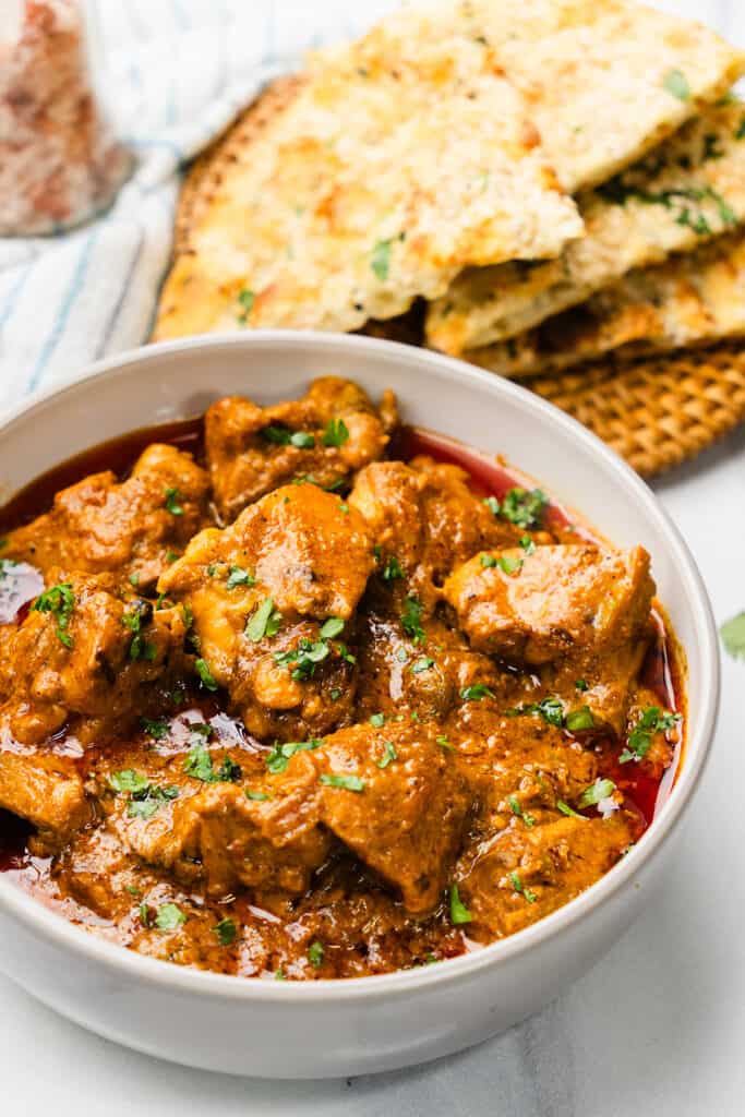 A bowl of Chicken Korma with cilantro on top, next to garlic naan on a woven placemat, with a striped cloth in the background.