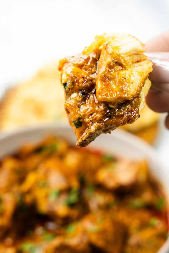 A piece of naan bread dipped in Chicken Korma, with a blurred bowl of the dish in the background.