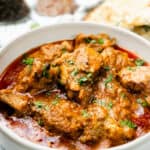 A bowl filled with Chicken Korma, garnished with fresh cilantro, sits beside garlic naan on a woven mat.