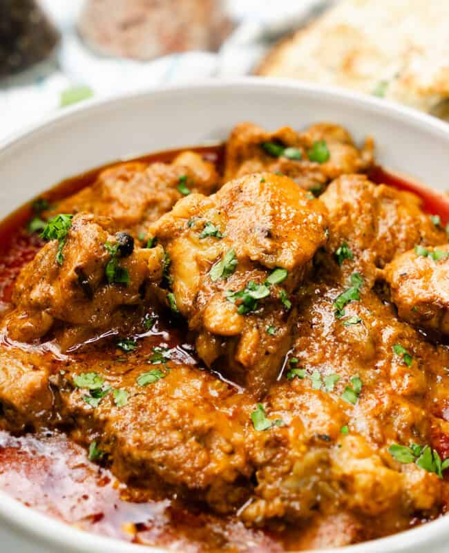 A bowl filled with Chicken Korma, garnished with fresh cilantro, sits beside garlic naan on a woven mat.