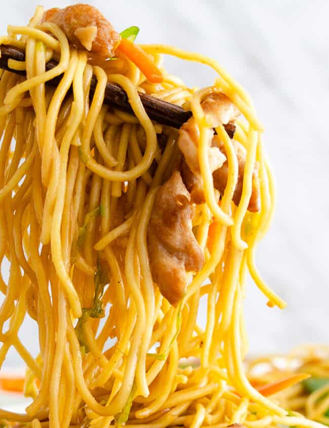 Chopsticks holding a chicken lo mein with noodles, vegetables, and chicken, set against a white background.