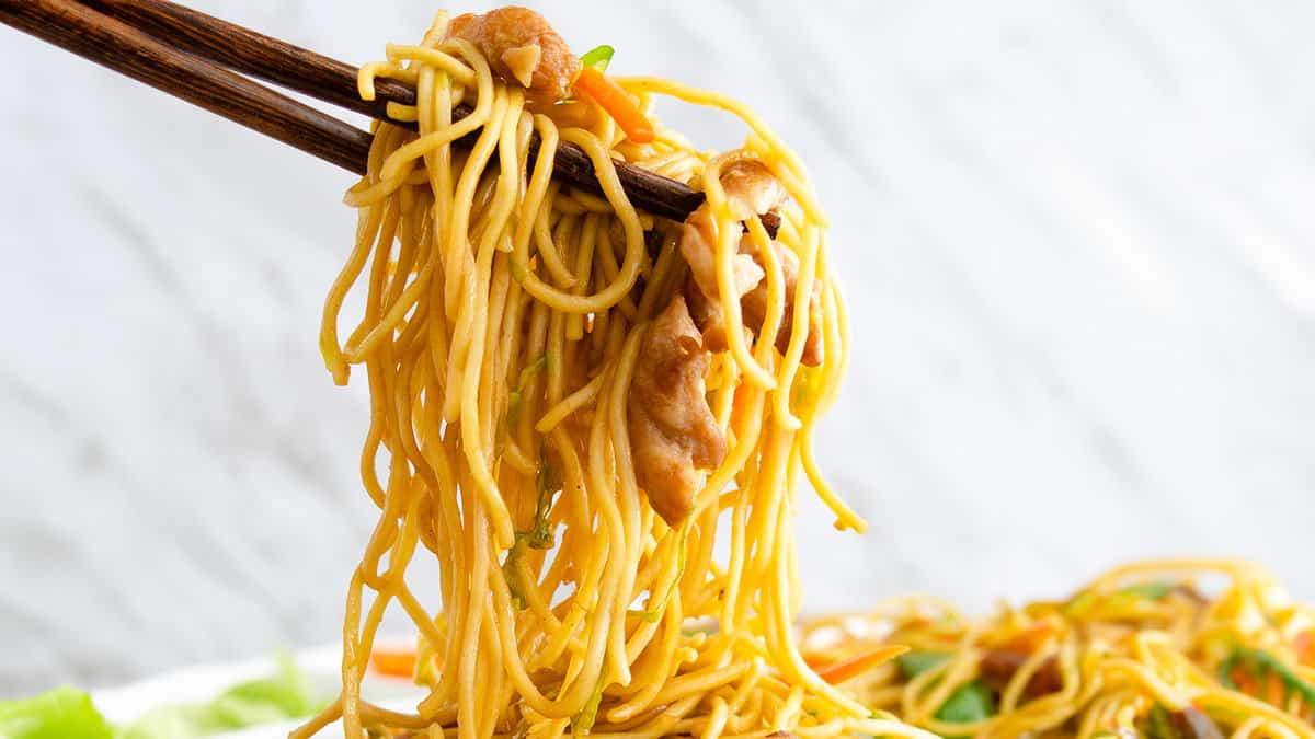 Chopsticks holding a chicken lo mein with noodles, vegetables, and chicken, set against a white background.