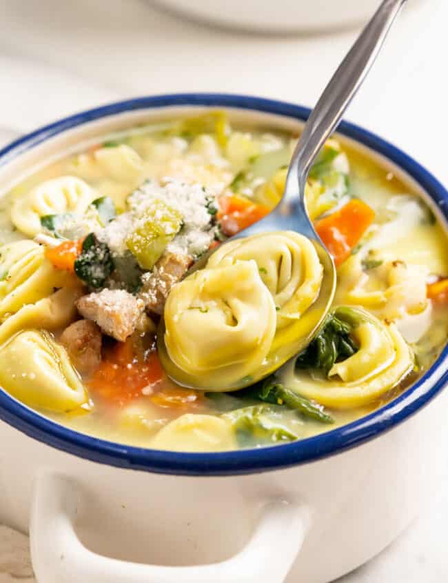 A close-up of a comforting Chicken Tortellini Soup in a white bowl with blue rim, with a spoon ready for enjoying.