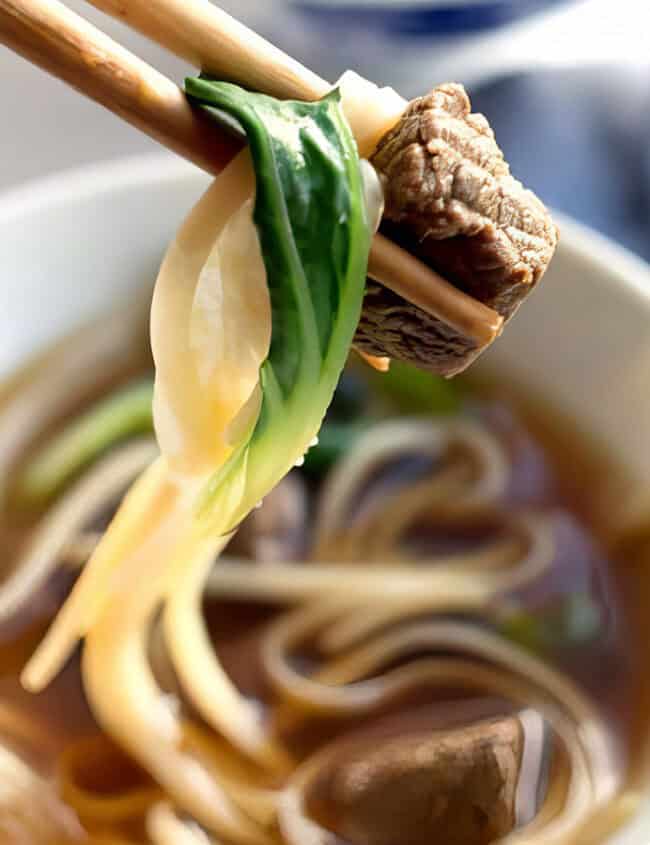 A white bowl containing beef noodle soup with a dark broth, slices of beef, white noodles, and green scallions on top.