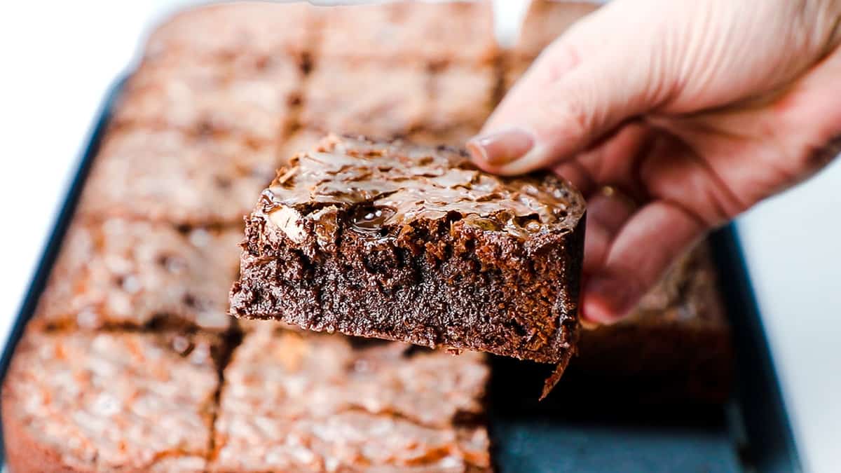 A woman's hand holding a fudgy brownie over a tray of Chocolate Chip Brownies.