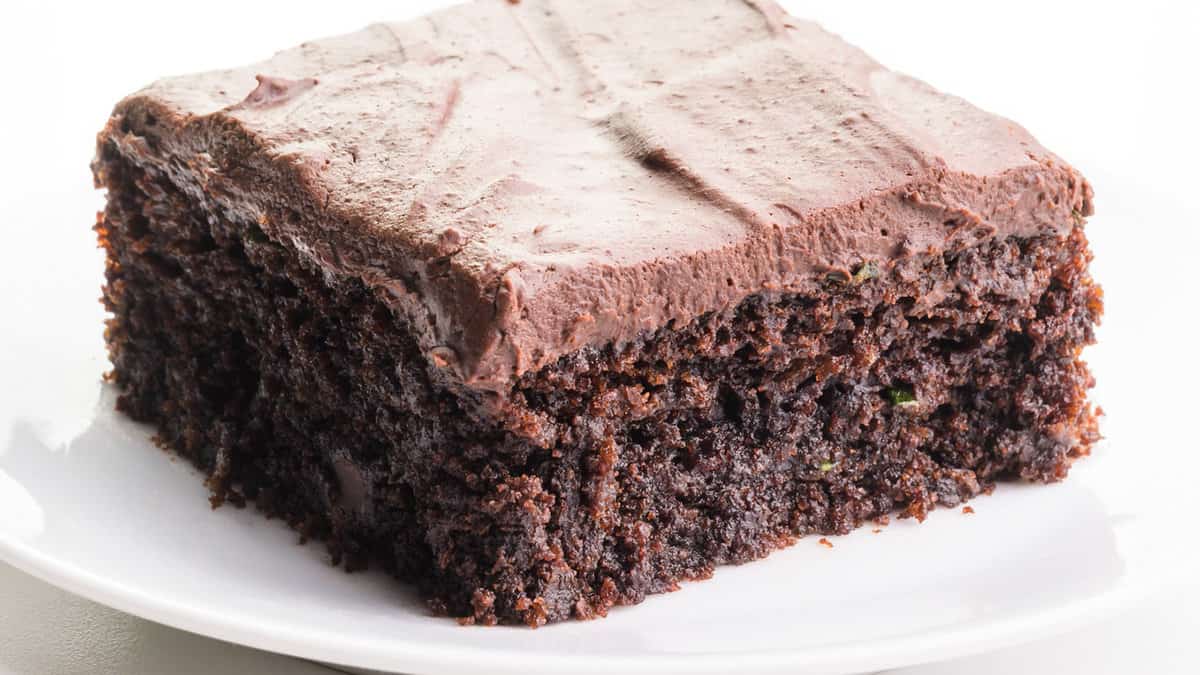 a slice of a frosted Vegan Chocolate Zucchini Cake on a white plate.