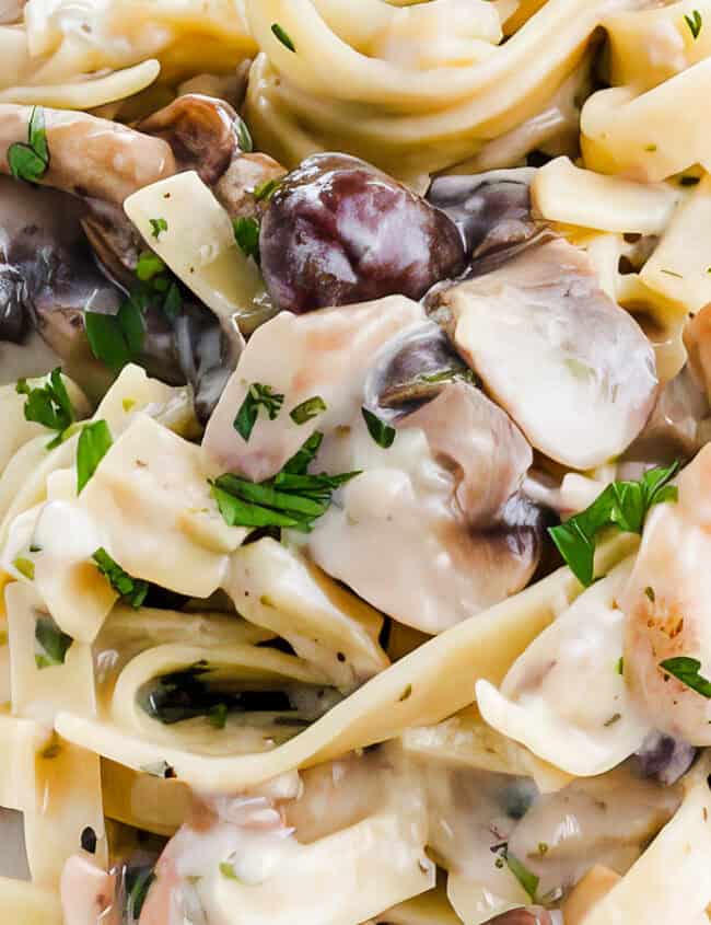 A close-up photo of a white plate overflowing with creamy tagliatelle pasta and sliced cremini mushrooms. A silver fork rests on the side of the plate.