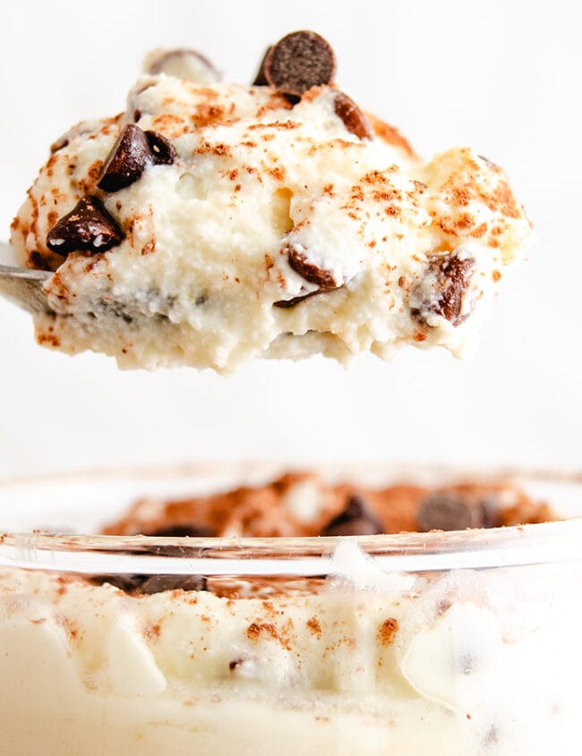 A spoon lifting a portion of creamy ricotta dessert sprinkled with chocolate chips and a dash of cinnamon, with more of the dessert visible in the glass below.