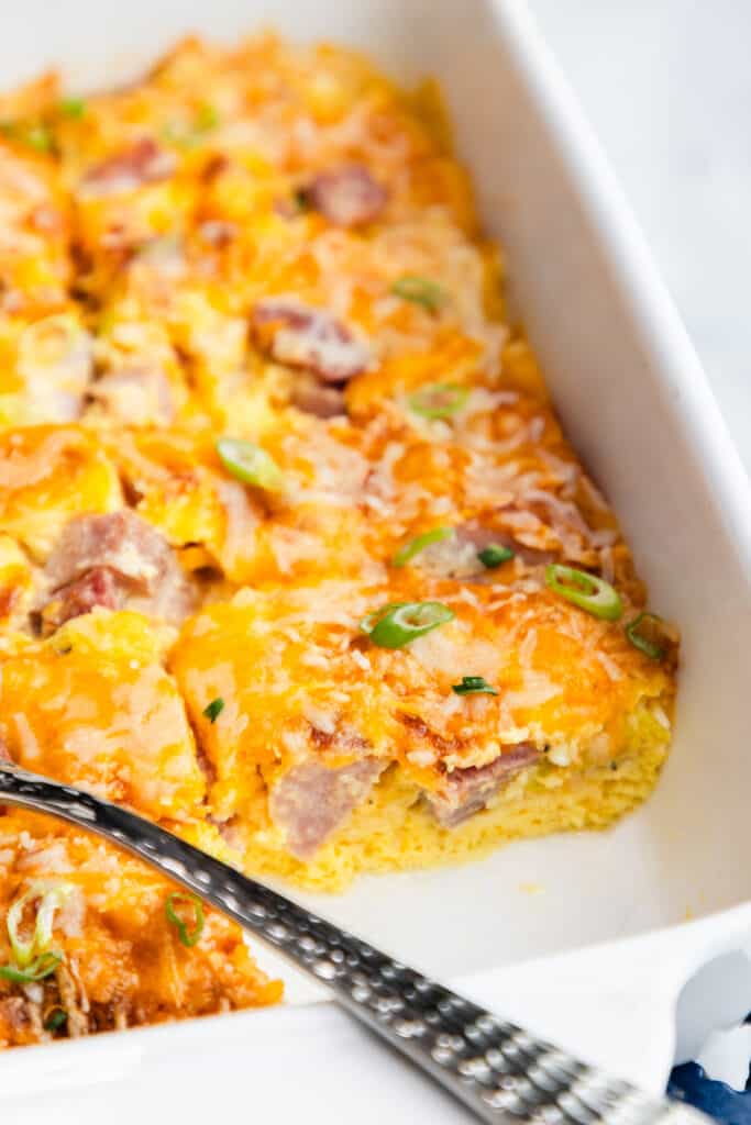 A spatula is used to scoop out a portion of ham and egg casserole from a white baking dish, topped with melted cheese and garnished with fresh green onions.