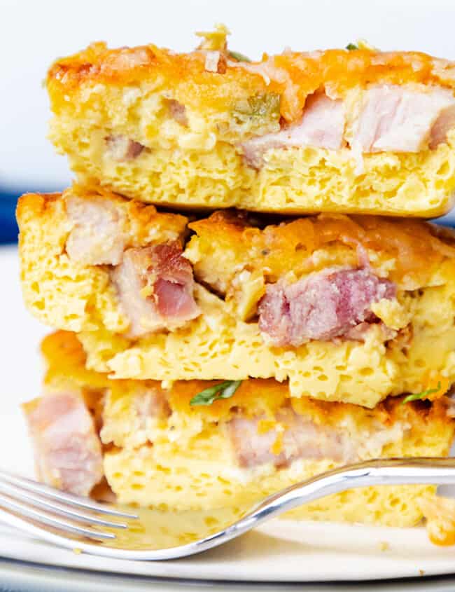 Three stacked slices of ham and egg casserole with melted cheese on a white plate, with a fork to the side.