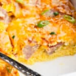 A spatula resting on a ham and egg casserole from a white baking dish, topped with melted cheese and garnished with fresh green onions.