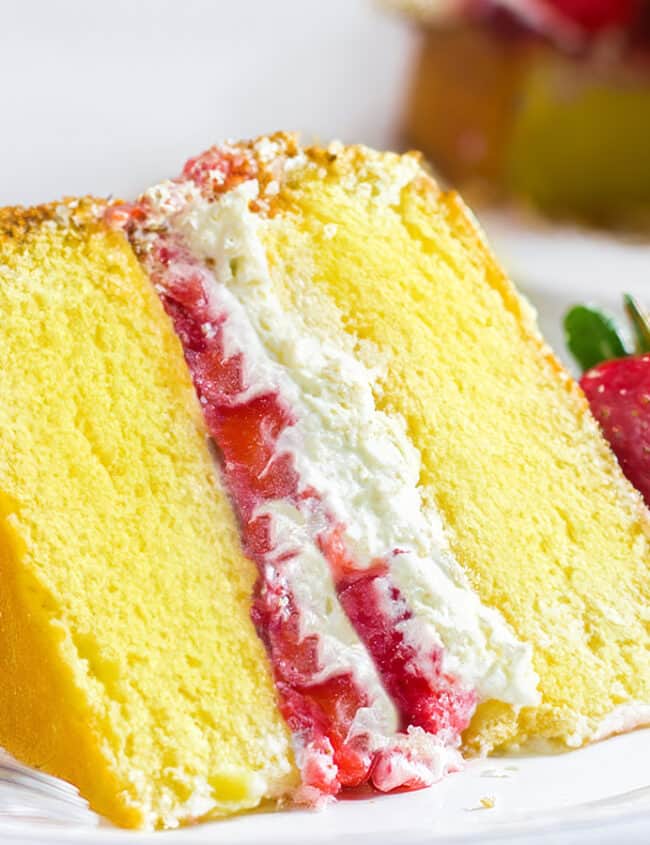 A slice of Lemon Sponge Cake filled with whipped cream and strawberries on a plate with the cake behind it.