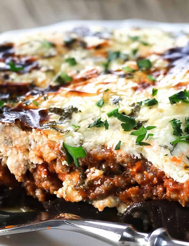 A slice of Moussaka on a white plate and a fork next to it.